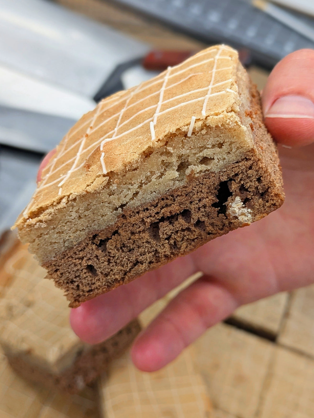 Hand holding a Buckabee Brownies Butterscotch Cinnamon Brookie, cut side to the camera, showing two baked layers — a pale golden cinnamon blondie on top and a fudgy butterscotch brownie below — with a crosshatch of white drizzle on the crackly top.