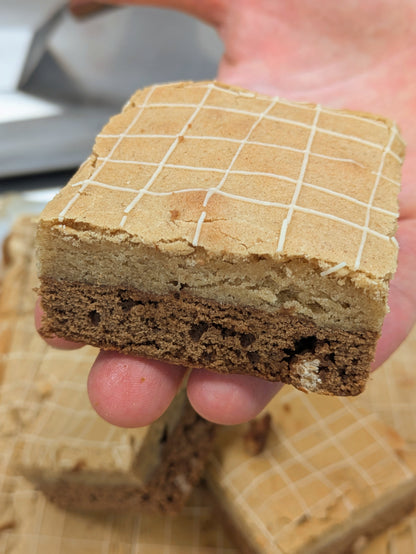 Hand holding a thick, square Butterscotch Cinnamon Brookie with the cut side forward, showing the golden cinnamon blondie top layer and butterscotch fudgy brownie bottom layer under a white drizzle grid.