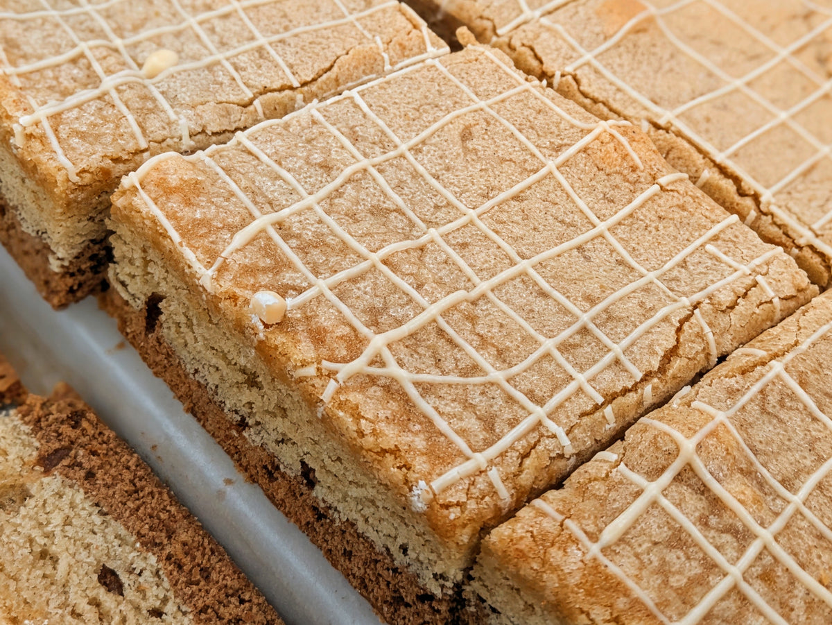 Overhead close-up of freshly cut Butterscotch Cinnamon Brookie lined up on a metal pan, their golden cinnamon-sugar tops finished with a white crosshatch drizzle.
