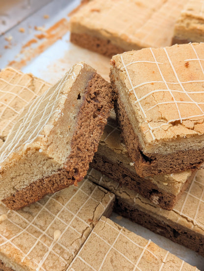 Angled close-up of stacked Butterscotch Cinnamon Brookies, one tipped on its side to reveal the two-layer cross-section — golden cinnamon blondie over butterscotch brownie — beside others showing their drizzled tops.