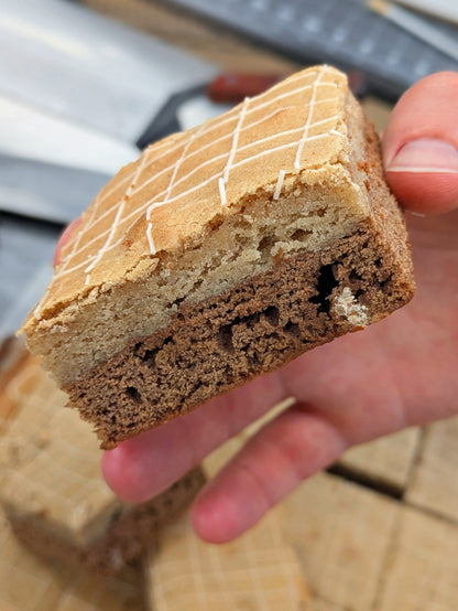 Hand holding a Buckabee Brownies Butterscotch Cinnamon Brookie, cut side to the camera, showing two baked layers — a pale golden cinnamon blondie on top and a fudgy butterscotch brownie below — with a crosshatch of white drizzle on the crackly top.