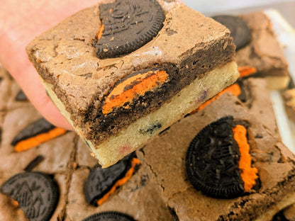A close-up of a hand holding a Cookies and Cream brownie topped with Halloween Oreo cookies and Confetti bottom layer and more cut brownies in the background.