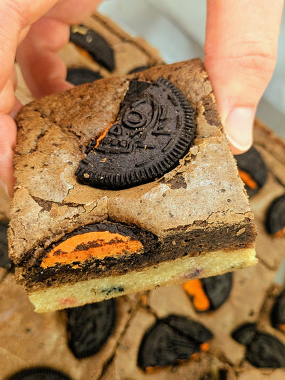 A close-up of a hand holding Cookies and Cream brownie topped with Halloween Oreo cookies showing orange cream filling, and Confetti Blondie on bottom layer.