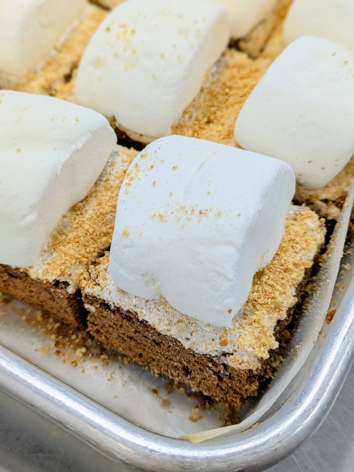 Close-up of a S'mores Brownie in a baking pan, topped with a large marshmallow and coated in crushed graham crackers over a fudgy chocolate brownie base