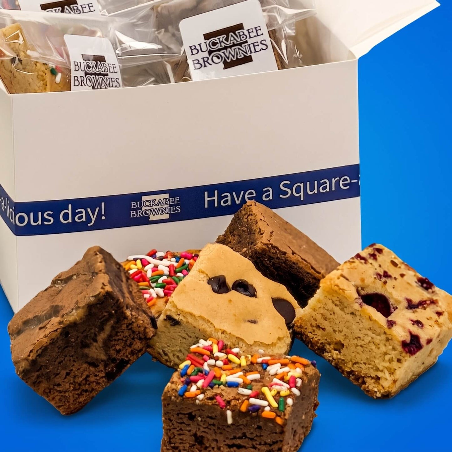 An open white gift box of Buckrabee Brownies' The Buckabee Experience Shipped Box with several brownie squares arranged in front on a bright blue background.