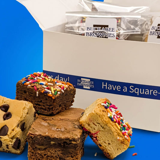 A box of Buckabee Brownies' Build Your Own Shipped Box with several brownie pieces arranged in front on a bright blue surface. The box is white with blue text and shows the Buckabee Brownies branding.