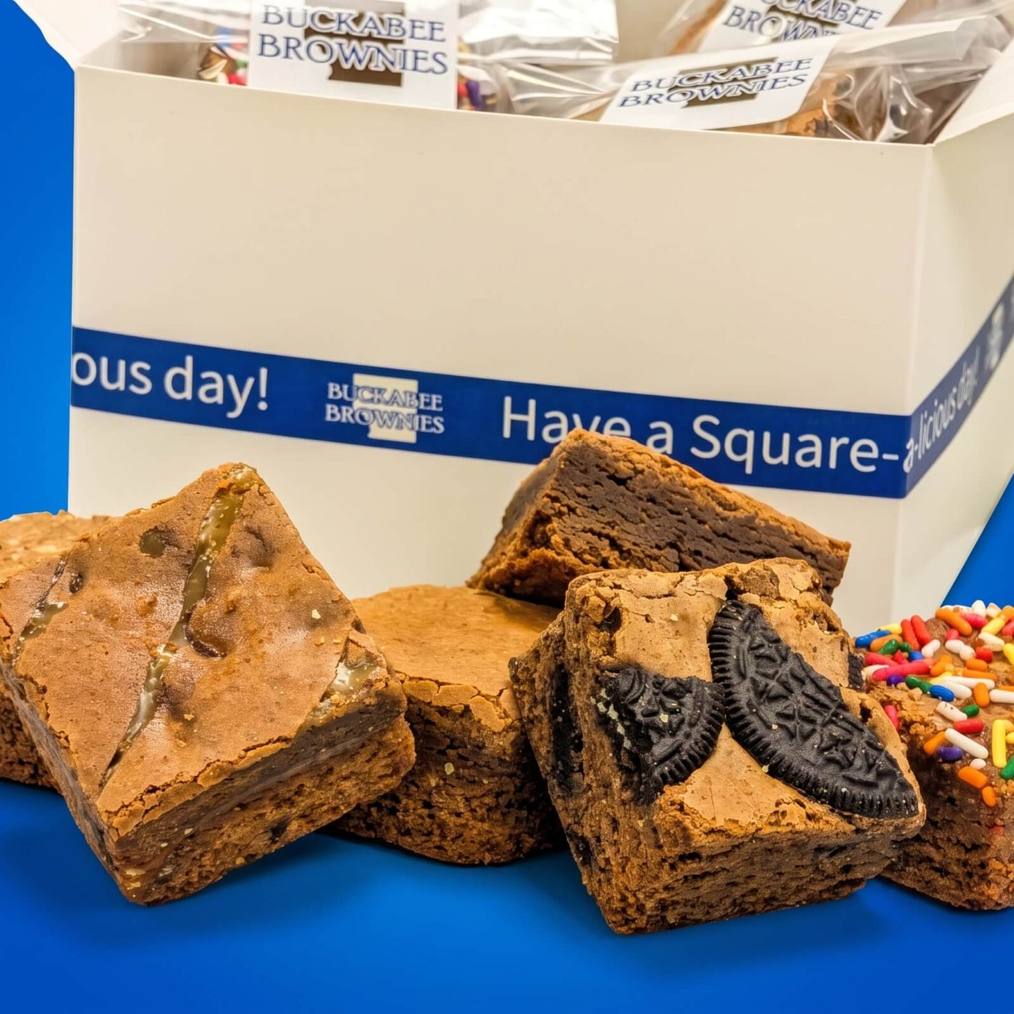 Buckabee Brownies' The Ultimate Chocolate Shipped Box filled with brownies, with several brownies displayed in front on a blue surface. Some brownies are topped with Oreo cookies, caramel and sprinkles.